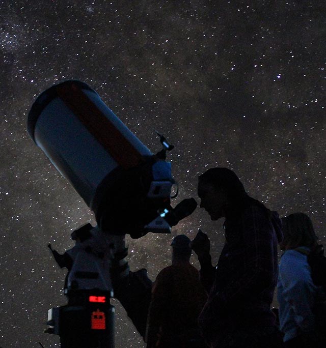 Students viewing the wonders of the night sky under a starry night sky, several people are taking turns looking through a large telescope.