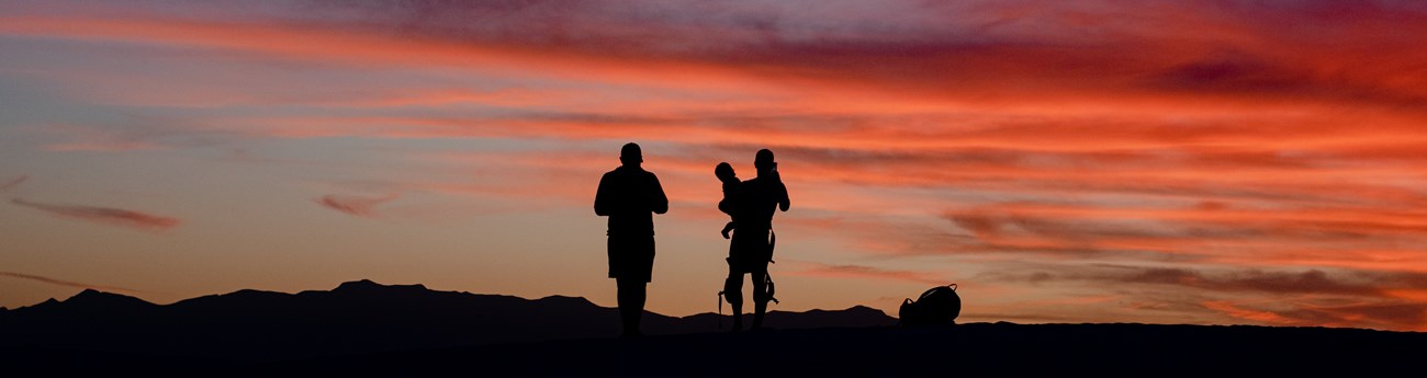 two visitors, one is holding a small child, silhouetted against a pink sunset sky