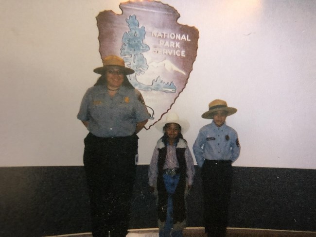 Diana Greymountain Park ranger stands next to two small children, one is wearing western cowboy wear and the other is wearing a park ranger uniform.