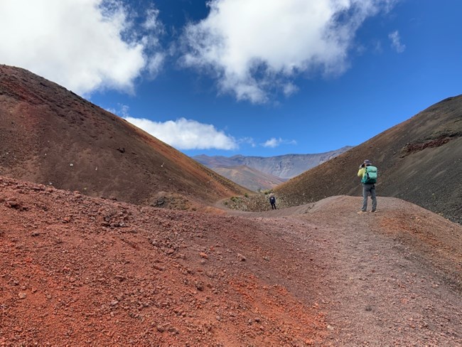 hiker in bright gear stands to take a picture between two cinder hills surrounded by colorful, orange cinder