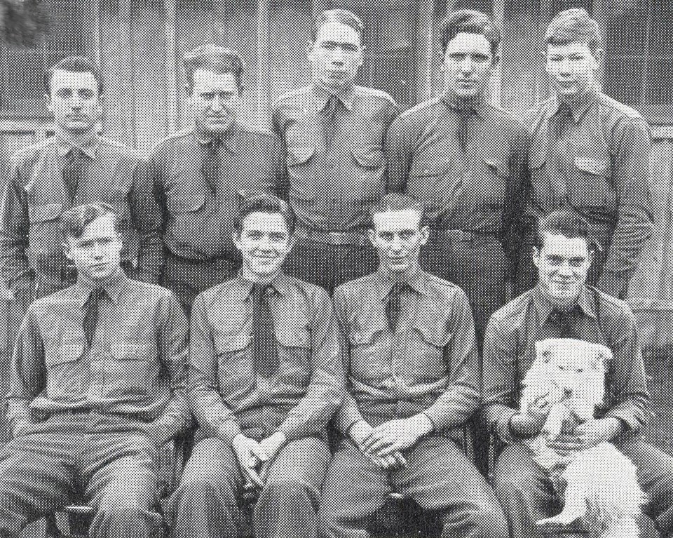 Black and white photo of young men wearing uniforms, standing and sitting. One seated man holds a white dog.