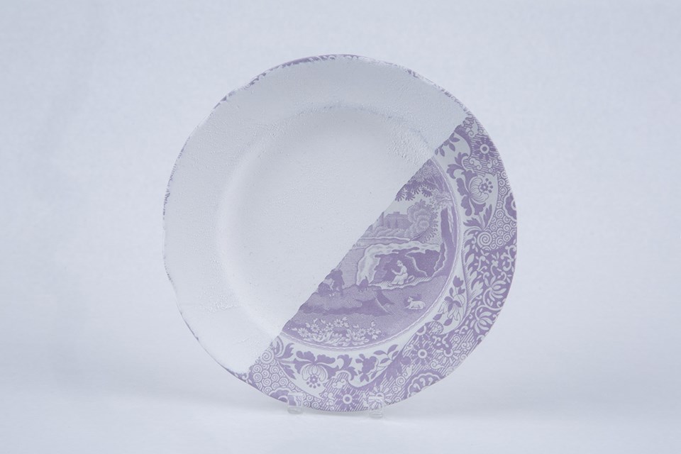 A small plate with a rural scene on it. The color of the print is light purple. Half of the plate has been dipped in white glaze.