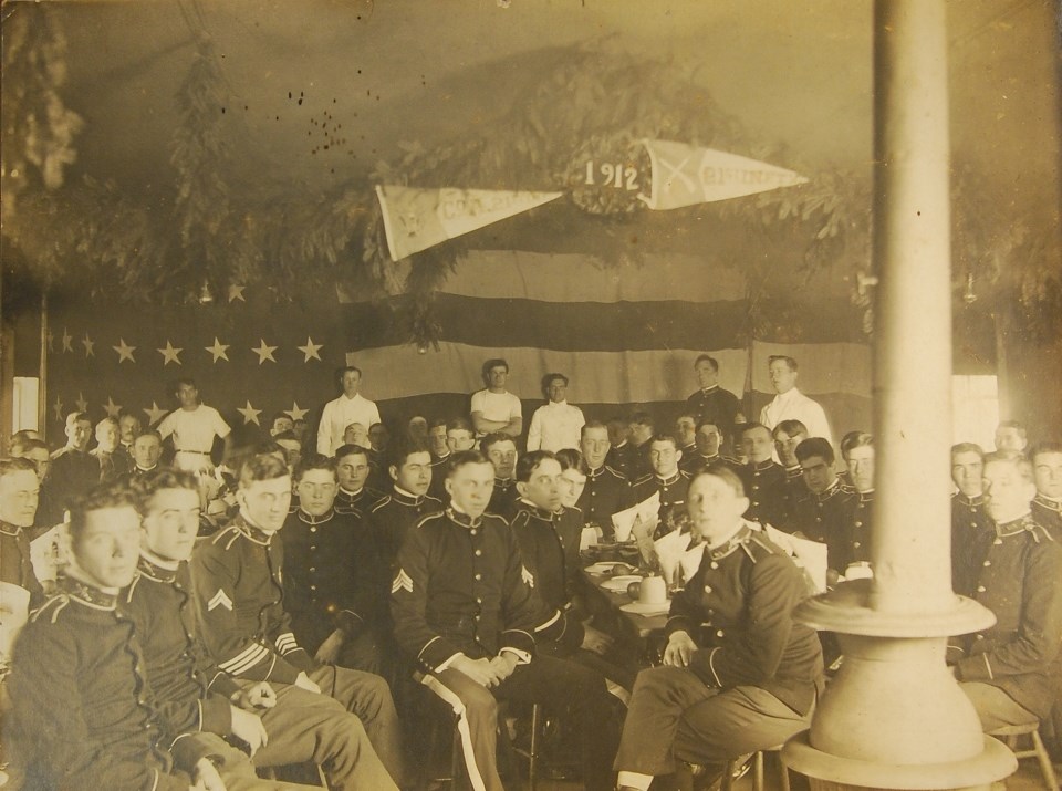 Photo of group of seated soldiers indoors. Behind them is a large American flag. Hanging above is Christmas greenery and banners that say 1912 and 21st Infantry.
