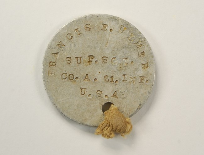 Round aluminum dog tag with engraved writing: Francis E. Ulmer, Sup. Sgt. Co. A. 21. Inf. U.S.A.