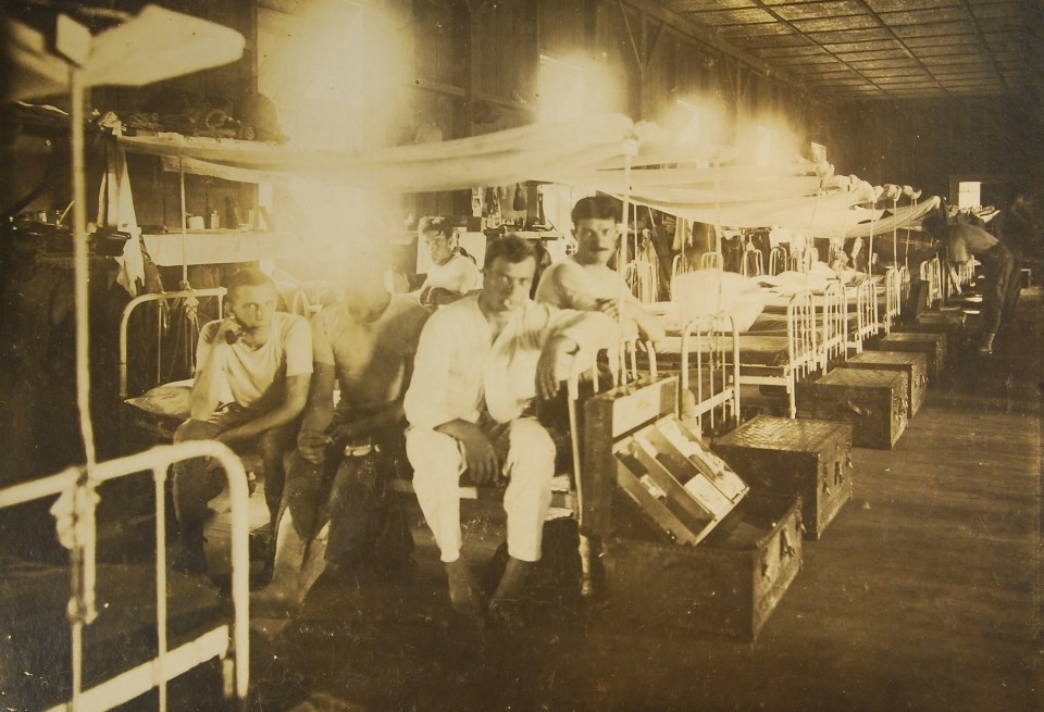In a barracks building with a row of beds, a group of men sit on a bed in the foreground dressed informally in shirts and slacks.