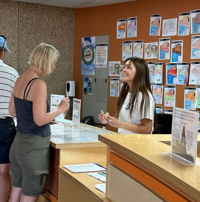 A Person stands behind a tall desk in a volunteer uniform talks with a memeber of the public.