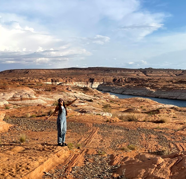 A person stands posing with their arms out on a red rocky surface. a lake is visable in the background.