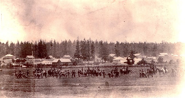 Soldiers, horses and cannons stand in a field. Behind them are barracks buildings.