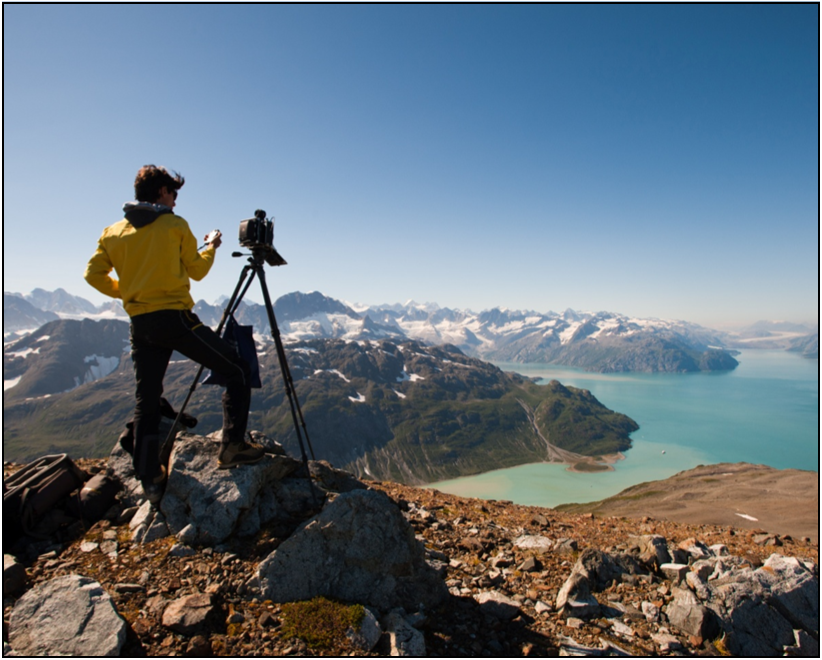 a person stands near a tripod with a camera on it balanced on a rocky cliff about fjords and mountains