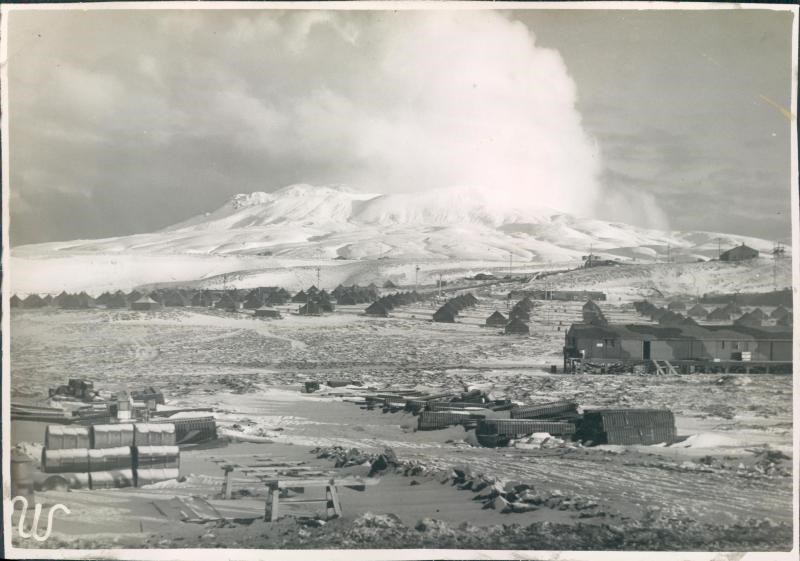 lt= Black and white photo of small tents and buildings with snowy mountain in background.