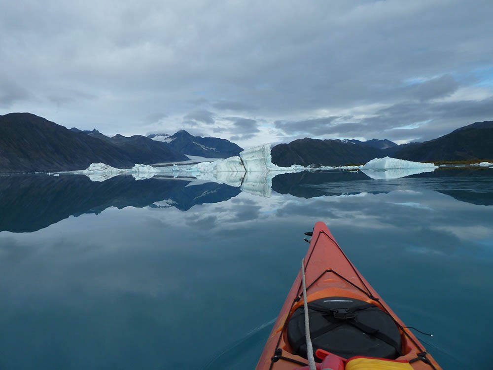 A kayak noses through a lake with icebergs and a glacier and mountains in the background.