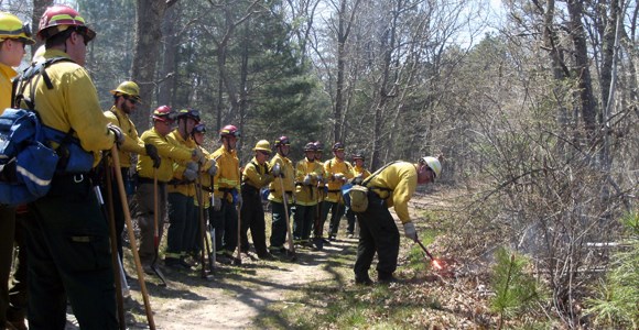 lt= A group of firefighters is gathered while the lead firefighter ignites grass with an ignition device.