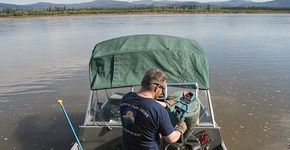 A man makes some adjustments to equipment on a boat along a river.