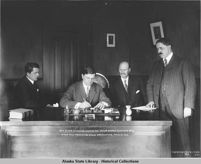 women 3 four men at a desk watching one of the group sign a document