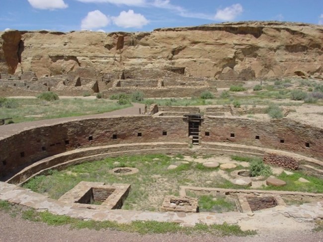 A subterranean sandstone circle known as a great kiva with tall canyon walls in the background.