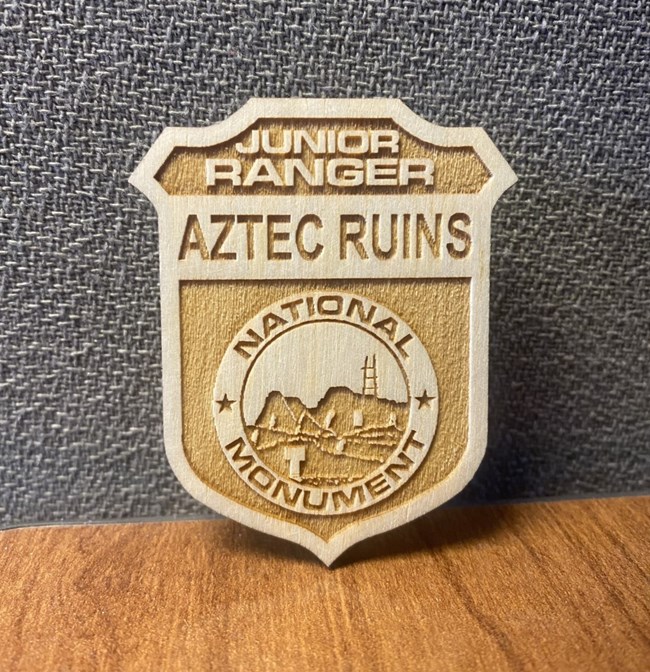 A wooden badge that says Junior Ranger Aztec Ruins National Monument.