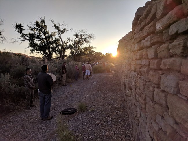 The sun rises adjacent to a long stone wall, with people and scraggly bushes nearby.