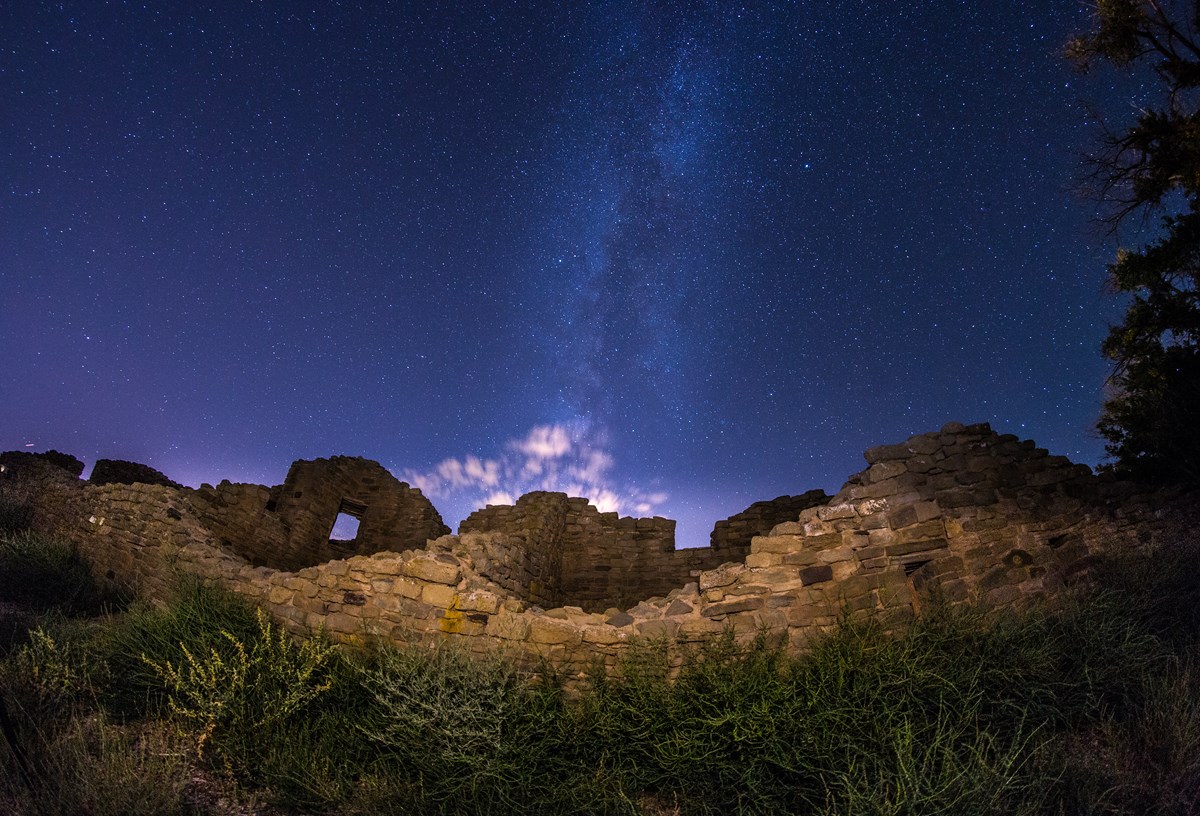 The milky way rises above stone ruins, with a burst of bright clouds and vibrant bushes in the foreground.