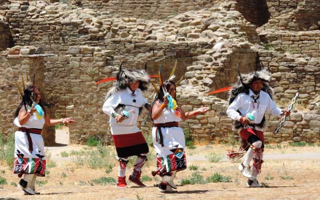 Tribal dancers in bright costumes in front of stone ruins::Pueblo dancers at Aztec Ruins