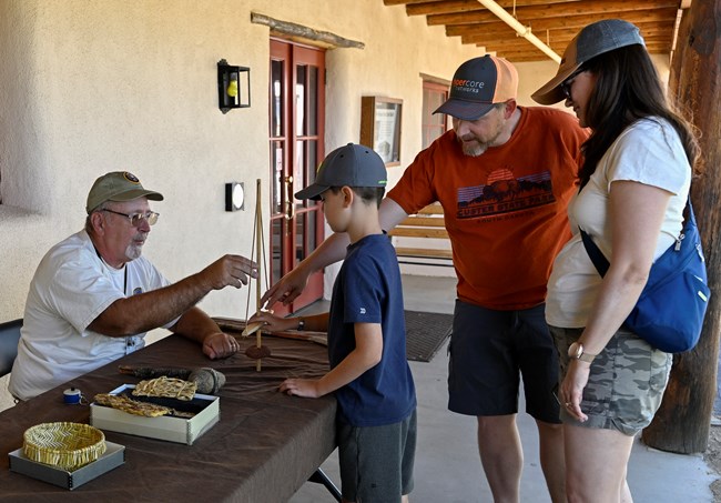 A volunteer sitting behind a table with replica items displayed talking to a family and showing them the items.