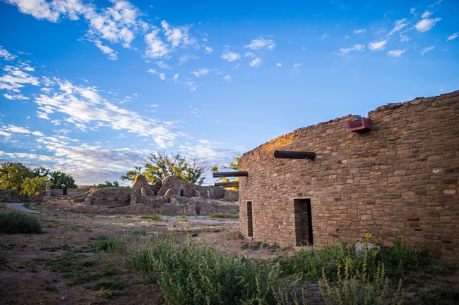 A circular sandstone structure in front of a series of sandstone walls. There are green grasses in the foreground.