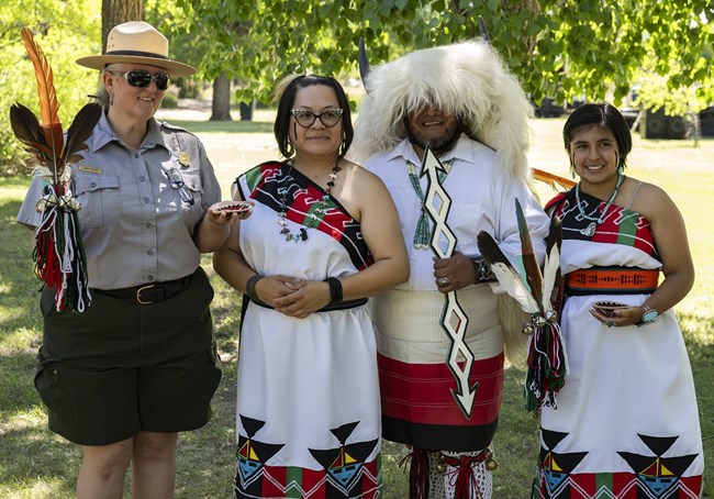 A ranger standing with a traditional Pueblo dance group. She is holding feathers and a woven basket and the dancers are wearing white, red, and green regalia.