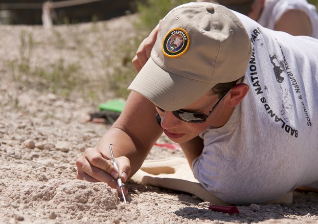 a man in sunglasses and a volunteer baseball cap uses a small pick to scrape at rock.