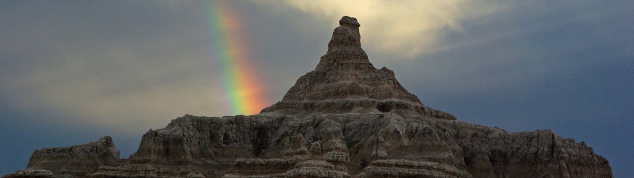 a rainbow disappears behind the single winding spire of a large butte.