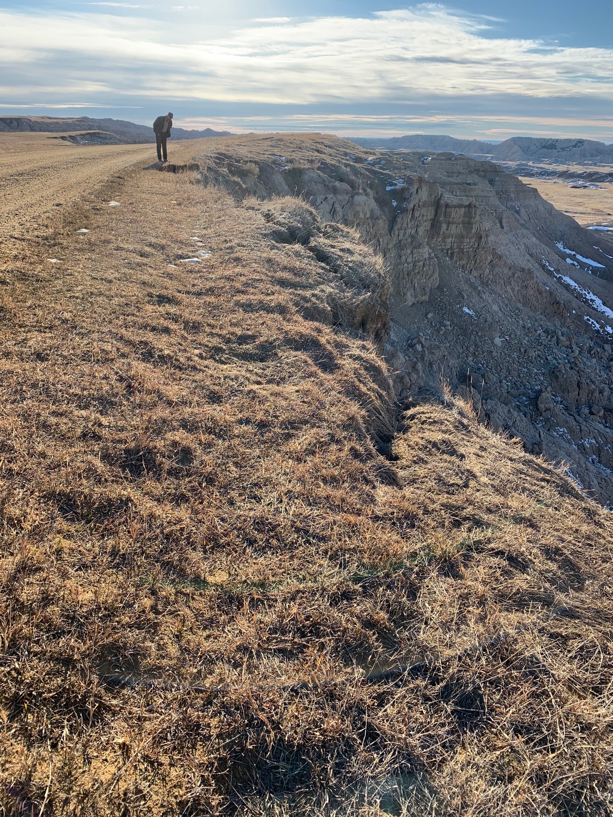 landslide at Sage Creek Rim Road Park Ranger standing at the edge of a dirt road. The shoulder of the road has eroded away and fallen down the cliff. Cracks are forming in the soil in the foregroud.