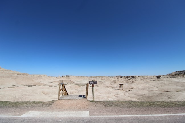 Trailhead for Castle Trail in a barren badlands landscape, with a small wooden footbridge and a sign marking the trail entrance, surrounded by pale, eroded rock formations under a clear blue sky.