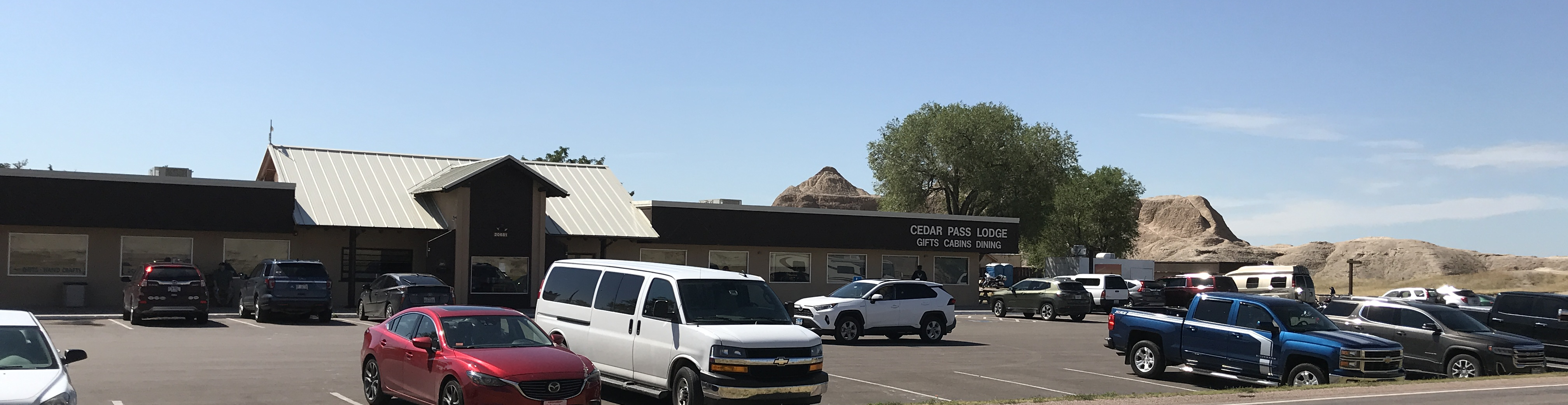 Several automobiles in front of tan building with sign reading "Cedar Pass Lodge, Gifts Cabins Dining."