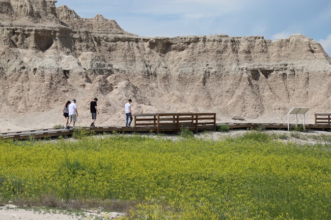 People walking along a board walk trail with informational signs along the way, and Badlands buttes in the background.