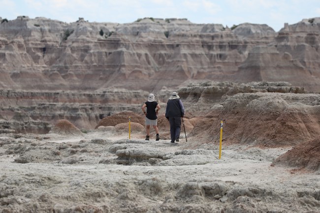 Two hikers walk along Door Trail in Badlands National Park, following a marked path through rugged badlands formations and layered rock cliffs in the background.