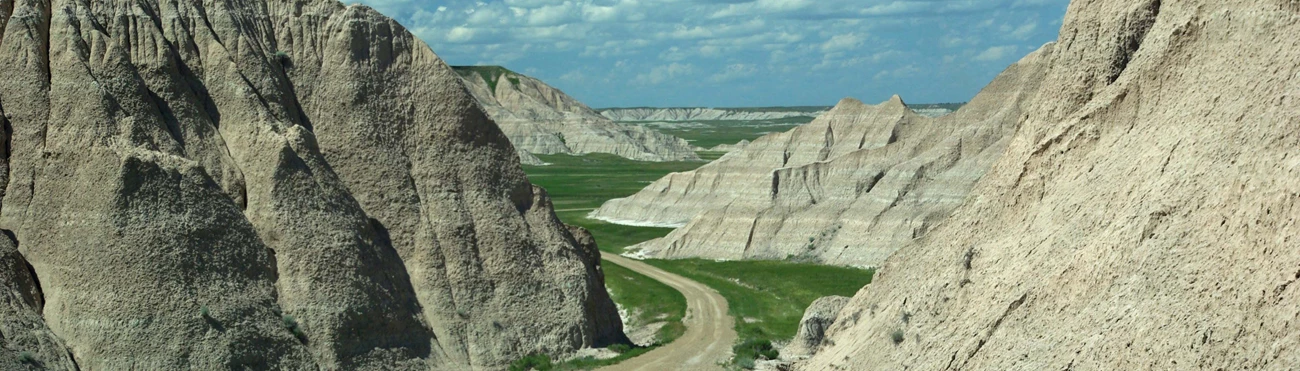 LarryMcAfee_SheepMountainRoad a dirt road winds between a pair of steep badlands buttes before disappearing behind the left butte, with a green grassy prairie in the distance.