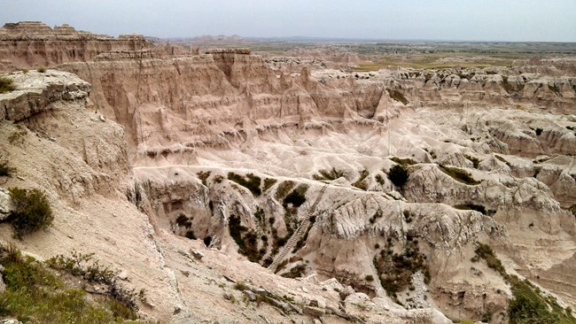 View from above the ladder on Notch Trail in Badlands National Park, looking out over rugged canyon walls, layered rock formations, and a sweeping valley below along the trail route.