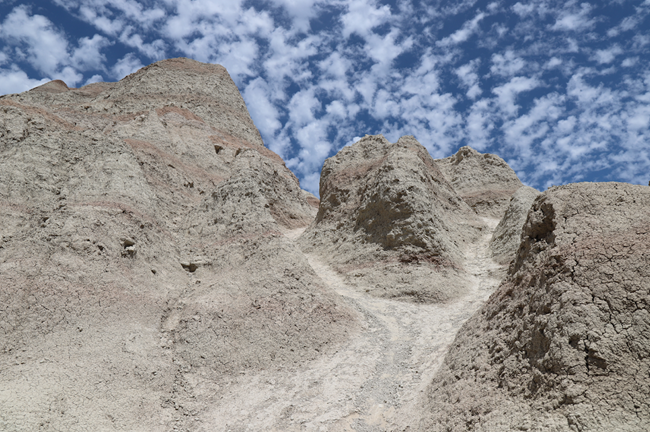 A worn trail forks in two directions headed up a steep butte with a cloudy blue sky overhead.