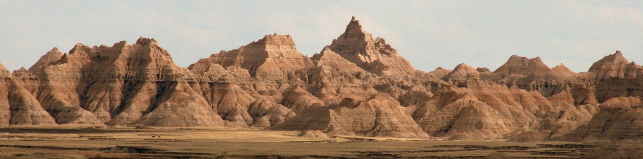 golden sunlight washes over the jagged peaks of badlands formations rising up and out of a dull green prairie.