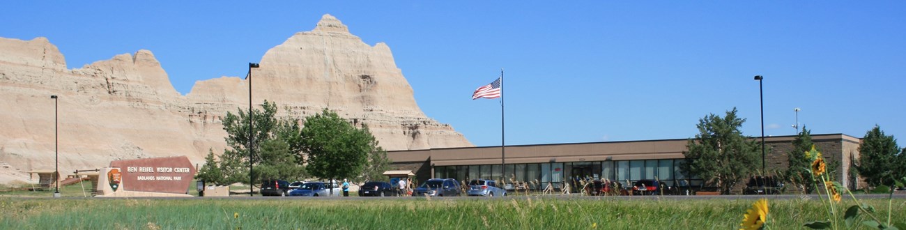a long, short tan building with a filled parking lot and sign sits in front of large badlands buttes.