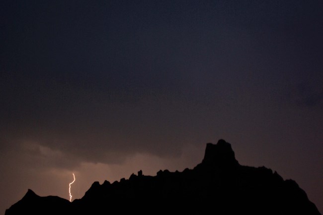 a crack of white lightning streaks out of a dark cloudy sky, silhouetted against the outline of badlands buttes.