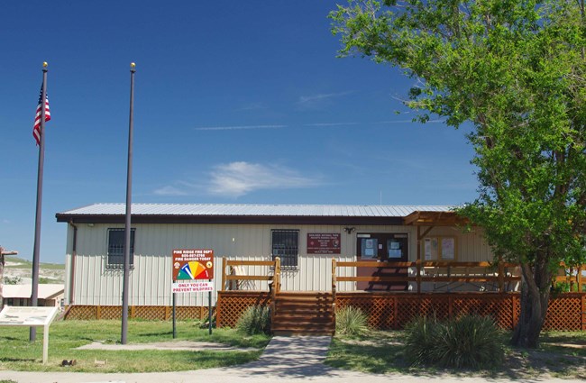 a squat tan building with a brown porch sits beyond green grass and a tree.