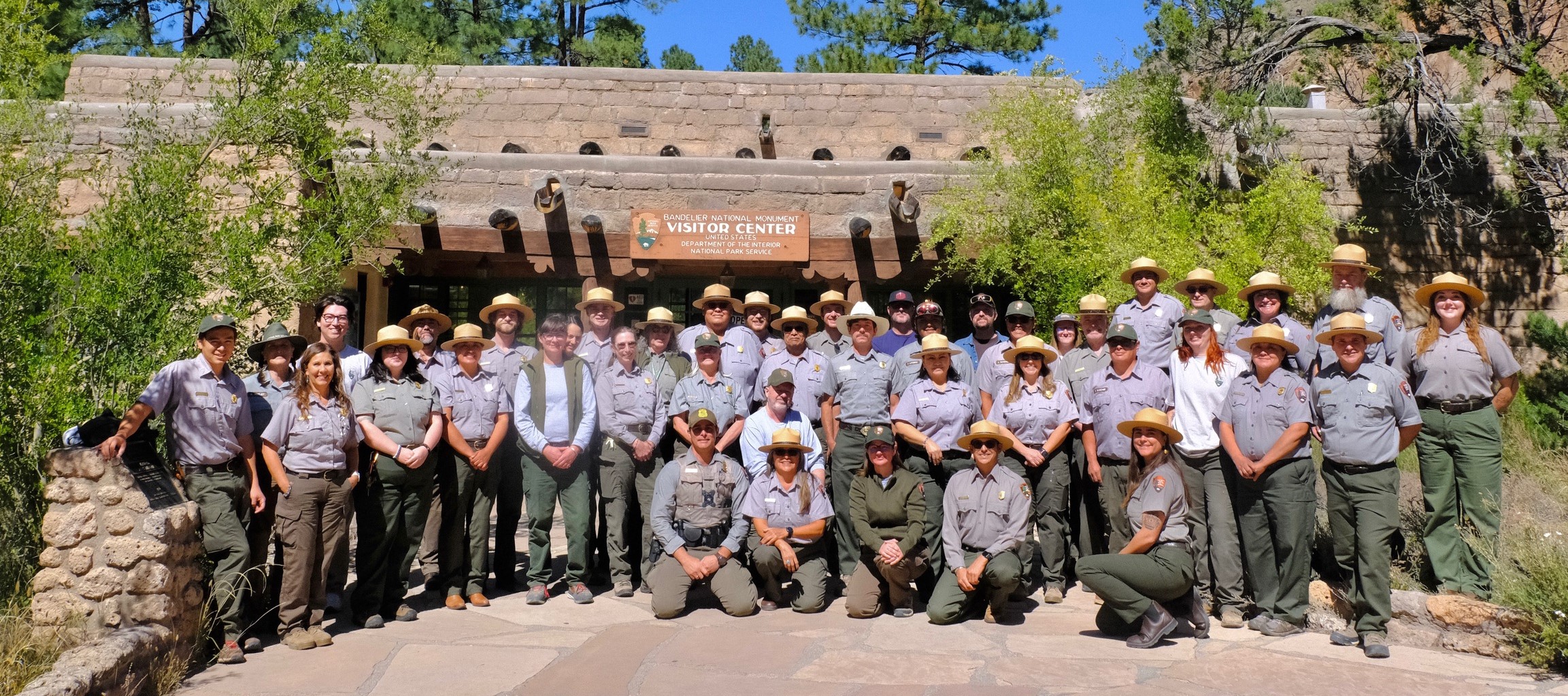 A large group of uniformed rangers stand smiling in front of the Bandelier Visitor Center