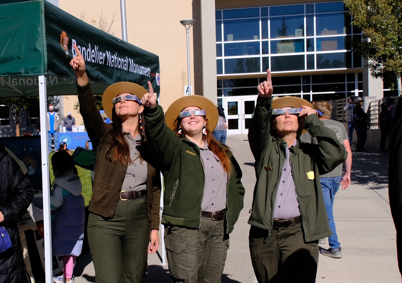 Three park rangers in uniform, wearing solar eclipse glasses, point at the sky and smile.