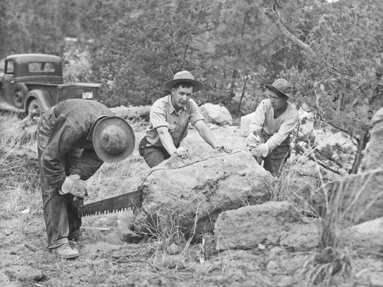 Three men with a handsaw cutting into a boulder.