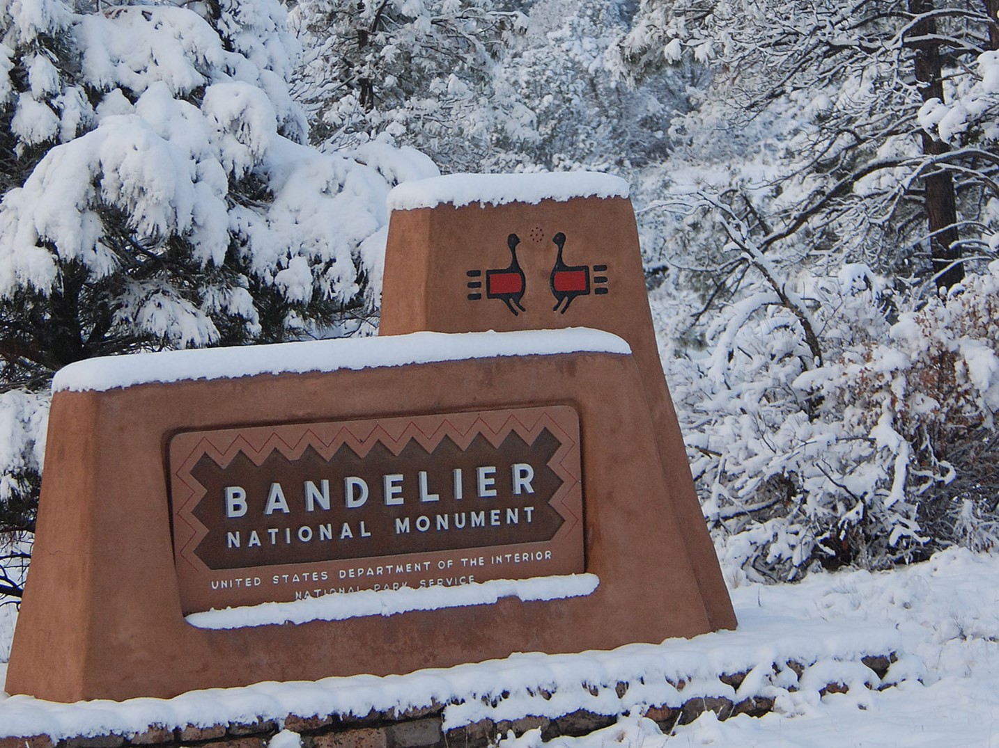 Bandelier National Monument will be closed on Thursday, January 4, 2024 due to expected heavy snow. snow lays across a brown sign with trees