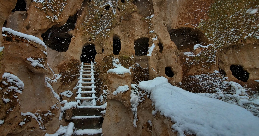White snow covers the ground and rocks. A ladder leads up to a hole in the side of the canyon.