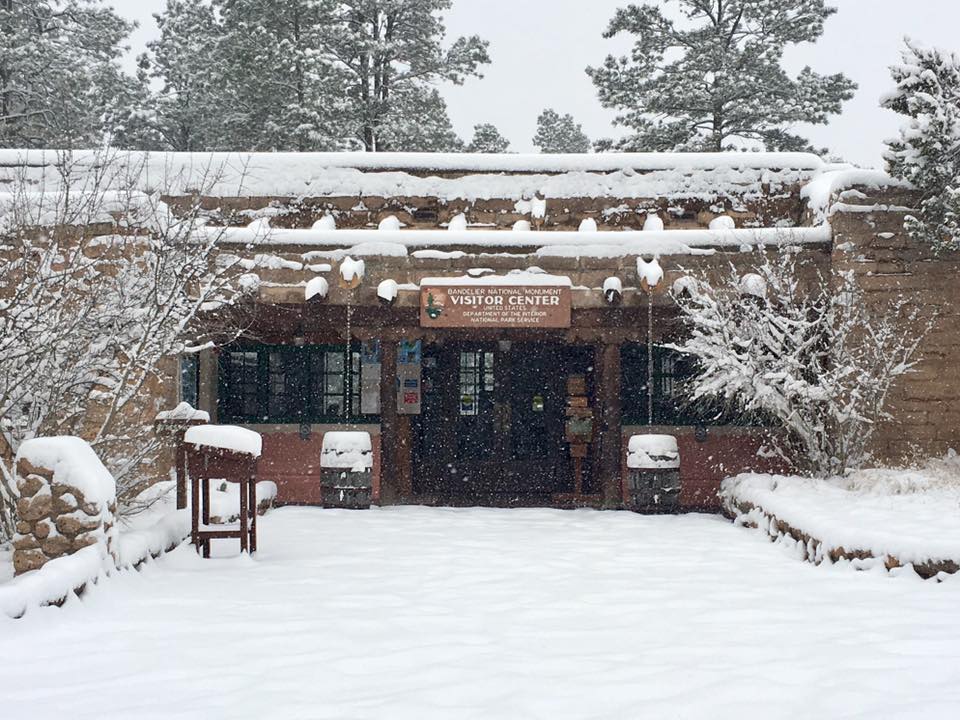 Bandelier National Monument will be closed on Friday, January 5, 2024 due to residual snow and park snow removal operations. White snow covers the ground and falls from the sky. A rustic building is covered with snow.