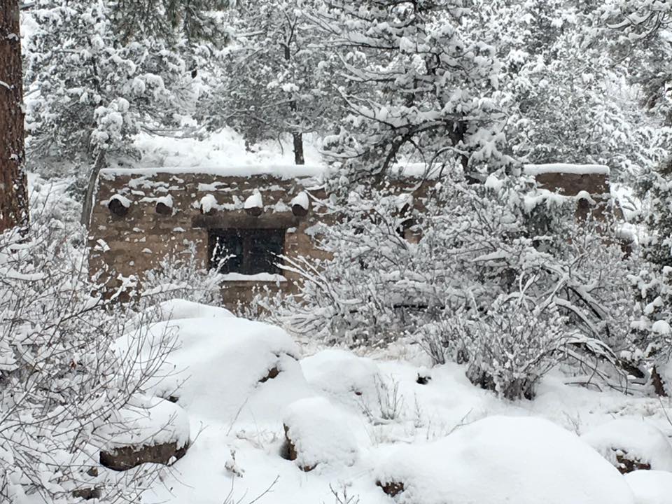 Bandelier National Monument will be closed on Thursday, November 30, 2023 due to winter weather conditions. White snow covers trees, bushes, and a rustic-looking building.
