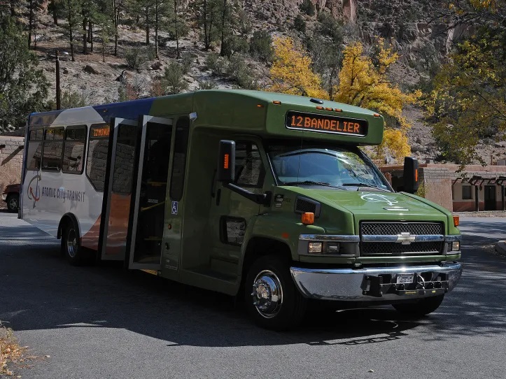 Bandelier Shuttle Bus A green, white, blue, and orange shuttle bus parked with its doors open. The shuttle's LED sign reads "12 Bandelier" in orange. The side of the bus reads "Atomic City Transit."