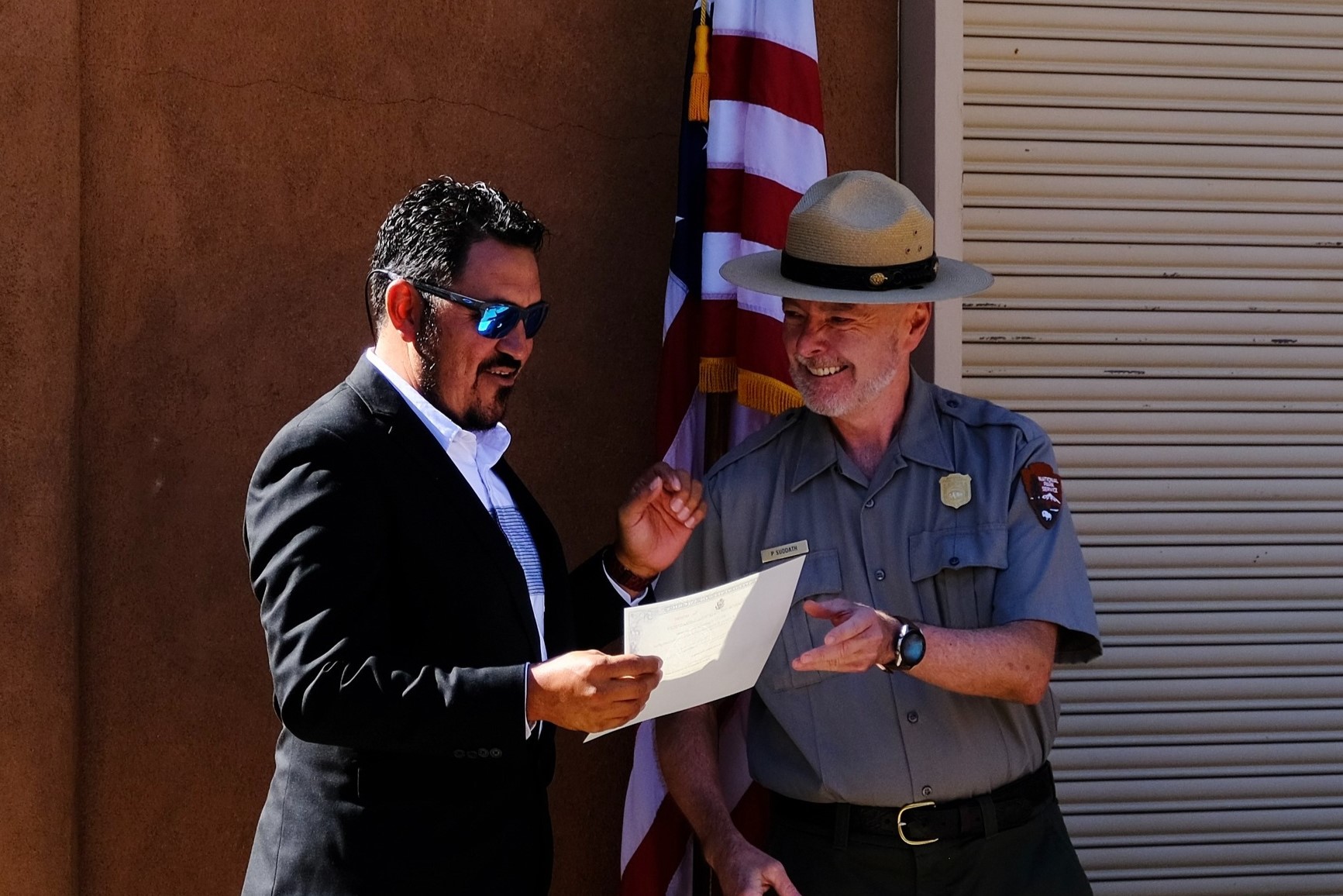 Bandelier National Monument, in partnership with U.S. Citizenship and Immigration Services, hosted a naturalization ceremony in honor of Constitution Day and Citizenship Day. A man in a suit holding a piece of paper standing beside a smiling park ranger. A United States flag is in the background.