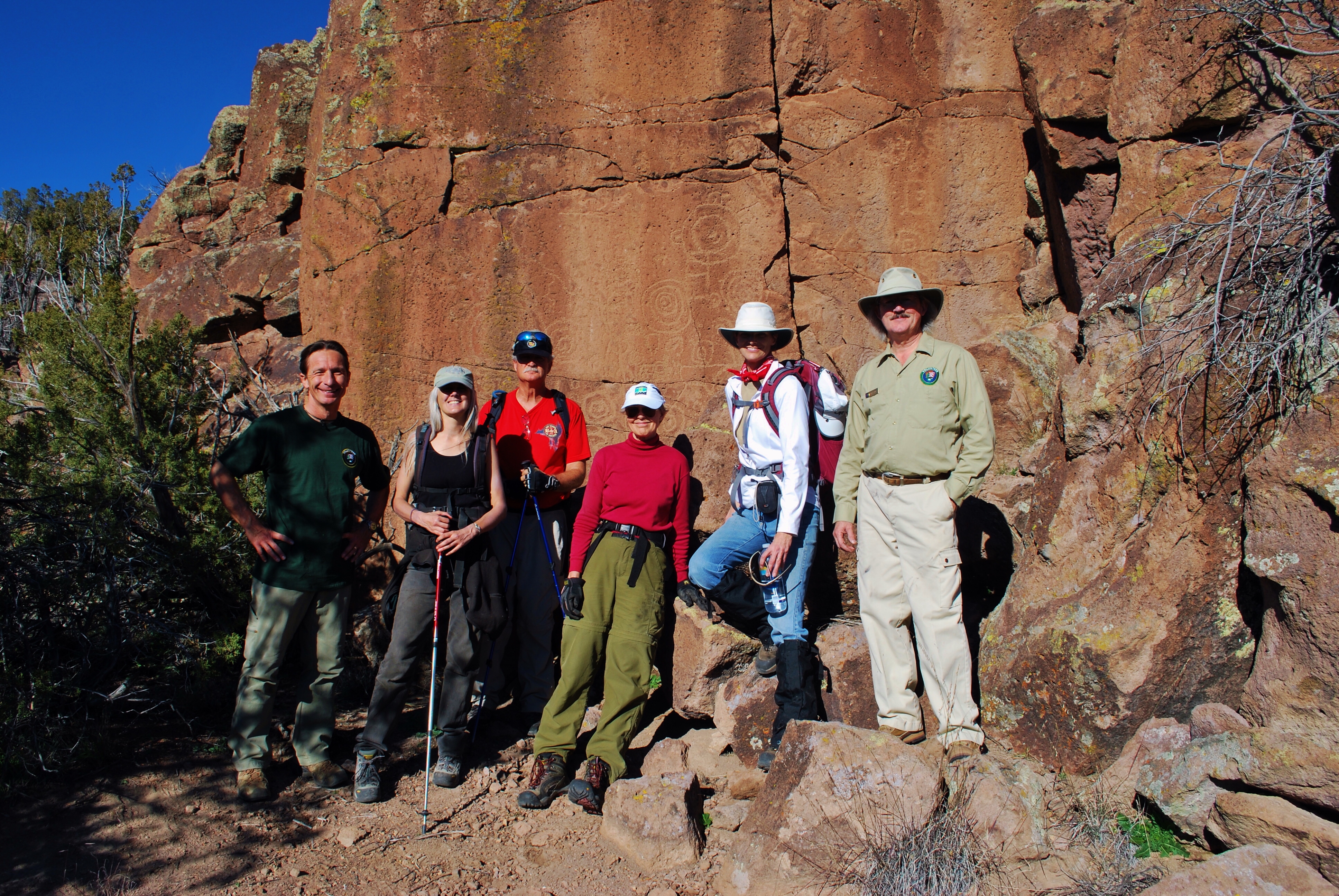 A group of six people standing in front of Ancestral Puebloan petroglyphs.
